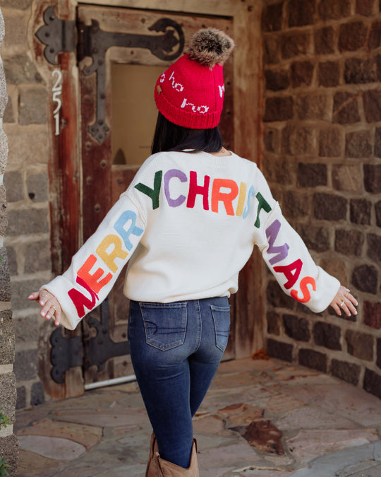 A person stands outdoors with arms outstretched, wearing a red beanie and a white sweater with MERRY CHRISTMAS in colorful letters on the back. Stone walls and a wooden door are in the background.