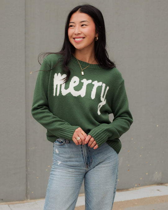 Woman wearing a beige knit beanie, brown sweater with WHISKEY WEATHER text, black leather pants, and gold necklace sits on a stool, looking to the side with one hand touching her hat.