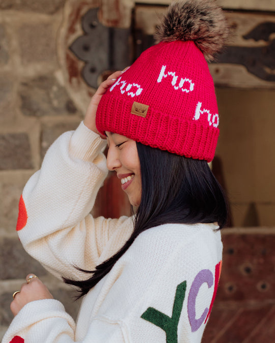 A smiling woman with long black hair wears a blue knit hat with a pom-pom and a white long-sleeve shirt, holding the sides of her hat against a dark background.