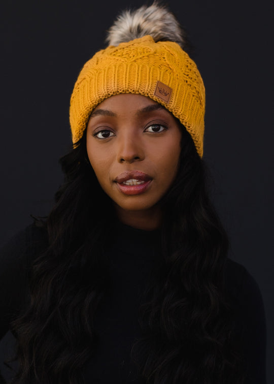 Woman wearing a yellow knit beanie with a fur pom-pom against a black background