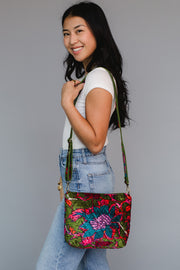 A woman in a white t-shirt and blue jeans smiles while posing with the Panache Apparel Collection Miki Floral Crossbody, which features an adjustable strap, against a plain gray background.