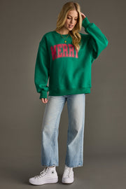 A woman with long blonde hair stands against a plain background wearing the Panache Apparel Collection's Merry Sweatshirt - Green with red lettering, light blue jeans, and white high-top sneakers, looking slightly downward.