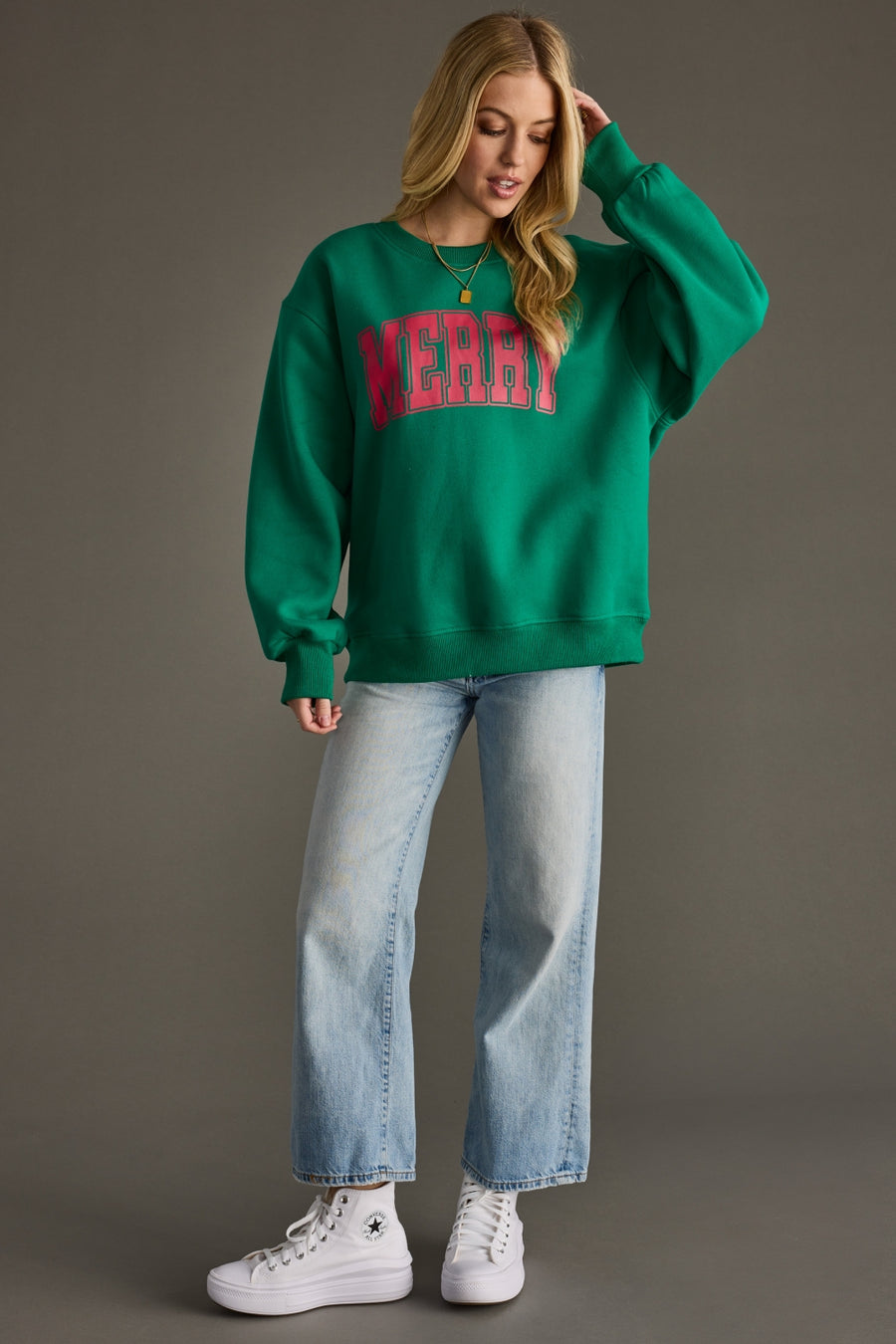 A woman with long blonde hair stands against a plain background wearing the Panache Apparel Collection's Merry Sweatshirt - Green with red lettering, light blue jeans, and white high-top sneakers, looking slightly downward.