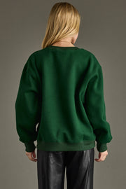 A person with long blonde hair, seen from behind, wears the Panache Apparel Collection Holly Jolly Sweatshirt in green and black leather pants, standing against a plain gray background.
