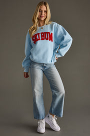 Woman wearing a light blue sweatshirt with 'Ski Bum' text, blue jeans, and white sneakers on a gray background
