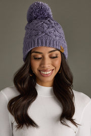 A woman with long wavy brown hair smiles while wearing the Flora Pom Hat by Panache Apparel Collection—a periwinkle knit hat with a decorative pom—along with a white ribbed top and gold hoop earrings, set against a gray background.