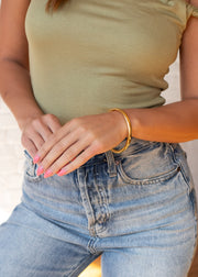 A person in a light green ribbed top and blue jeans stands with folded hands, displaying pink-painted nails and the Luxe Gold Thin Bangle by panacheaccessories. The background features a white textured wall.