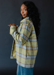 A woman with long wavy hair wears the Darby Plaid Jacket by panacheaccessories—a fully lined green and blue plaid piece with button closure—paired with blue jeans, standing sideways against a dark background and smiling gently.
