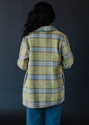 A person with long, wavy dark hair faces away, wearing the panacheaccessories Darby Plaid Jacket in yellow with button closure and blue jeans against a dark background.