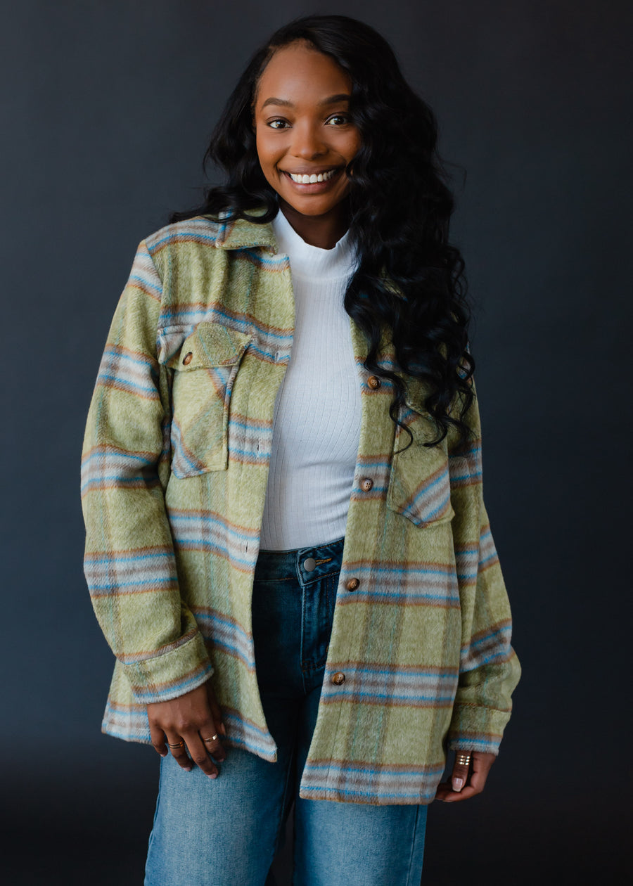 A smiling woman with long wavy hair models the panacheaccessories Darby Plaid Jacket with button closure, layered over a white top and blue jeans, set against a dark background.