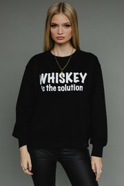 A woman with long blonde hair models the Panache Apparel Collection Whiskey Is The Solution Sweatshirt, paired with black pants and a gold chain necklace, posing against a plain gray background.
