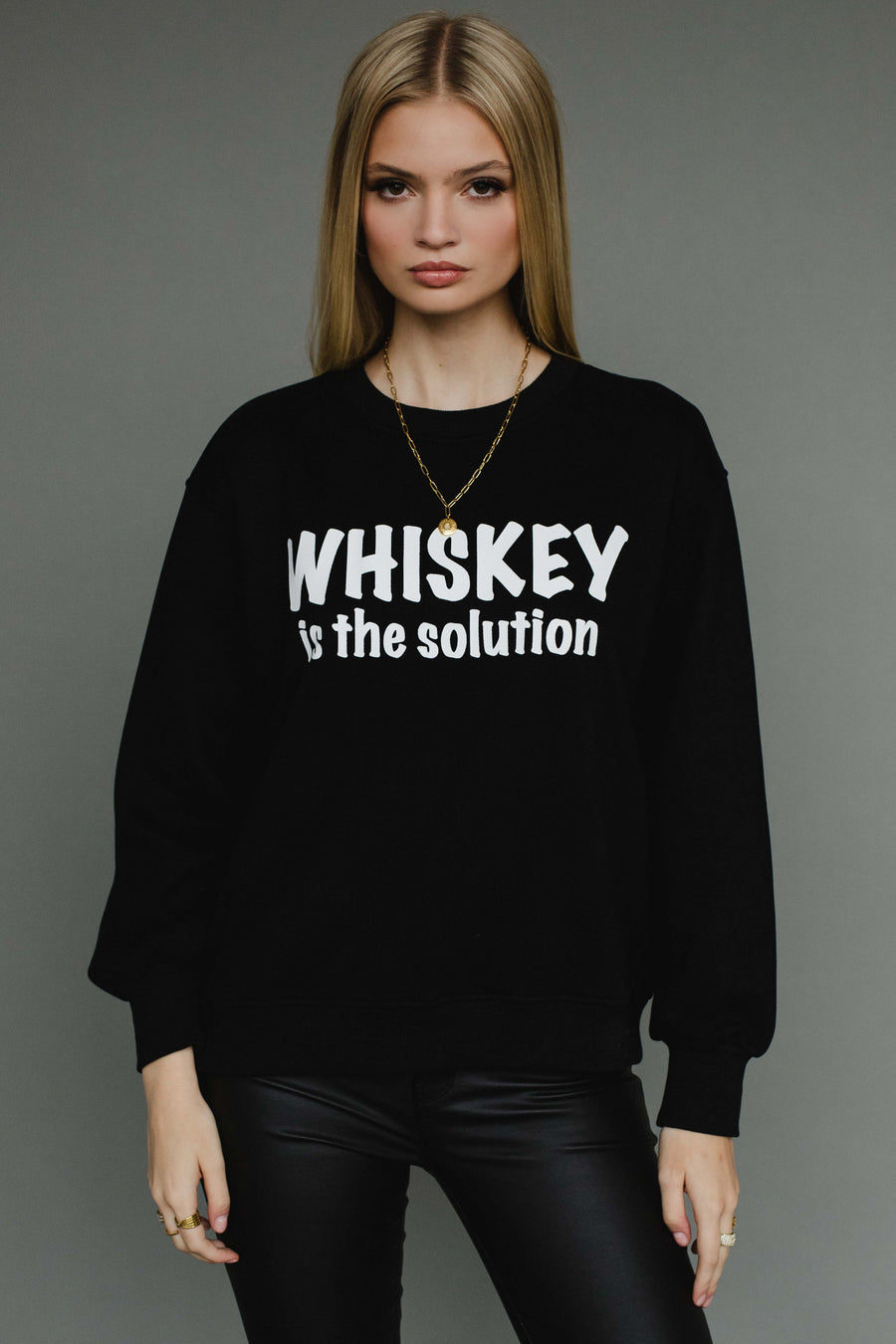 A woman with long blonde hair models the Panache Apparel Collection Whiskey Is The Solution Sweatshirt, paired with black pants and a gold chain necklace, posing against a plain gray background.