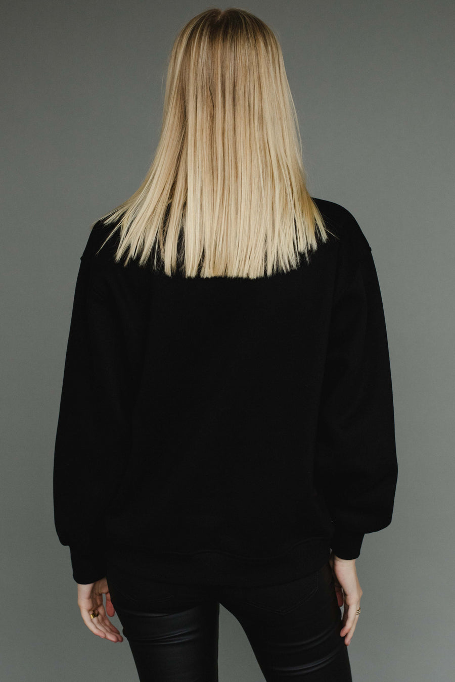A person with straight blonde hair stands with their back to the camera, wearing Panache Apparel Collection's black "Whiskey Is The Solution Sweatshirt" and matching pants against a plain gray background.