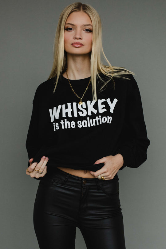 A woman with long blonde hair wears the Panache Apparel Collection's Whiskey Is The Solution Sweatshirt, styled with layered necklaces, rings, and black leather pants against a plain gray background.