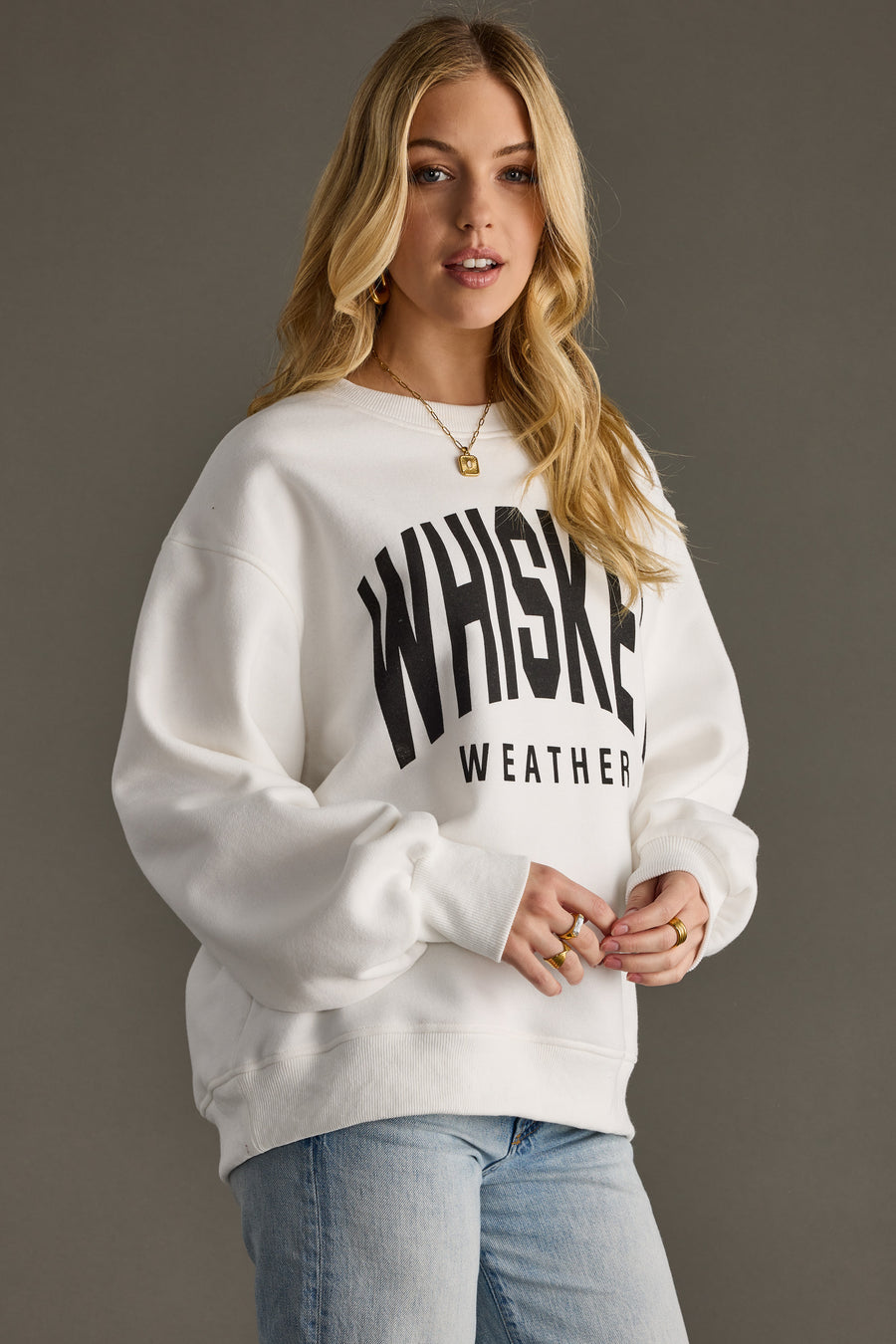 A woman with long blonde hair models the oversized Whiskey Weather Sweatshirt - White by Panache Apparel Collection, paired with light blue jeans against a plain gray background.