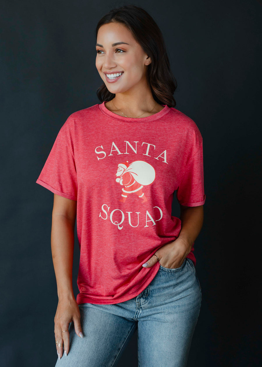 A woman smiles in the panacheaccessories Santa Squad Tee, a vintage red graphic t-shirt, paired with light blue jeans against a dark background.