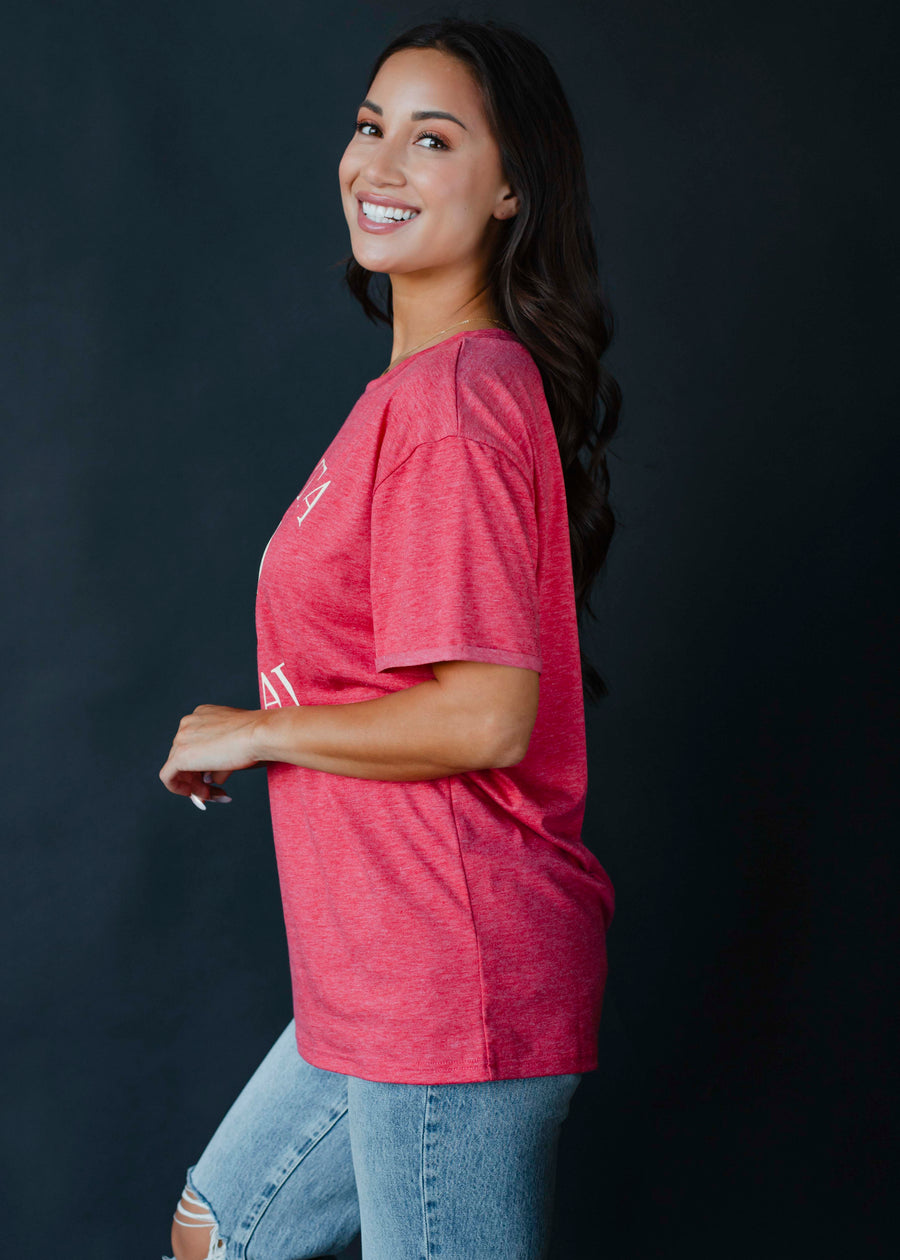 A smiling woman with long dark hair stands sideways against a plain dark background, wearing the oversized fit vintage red Santa Squad Tee by panacheaccessories and ripped light blue jeans.