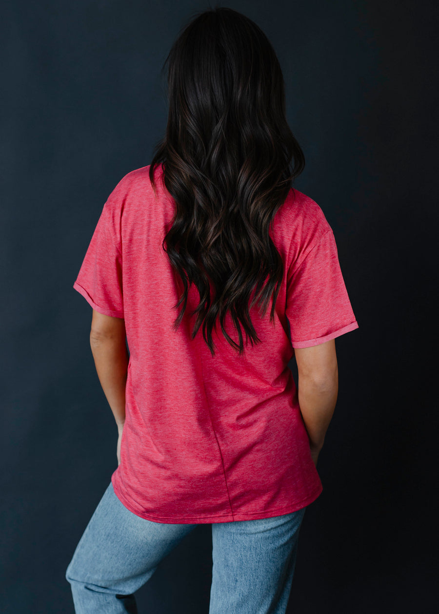 A person with long, wavy dark hair stands facing away, wearing the oversized vintage red Santa Squad Tee by panacheaccessories and light blue jeans against a plain dark background.