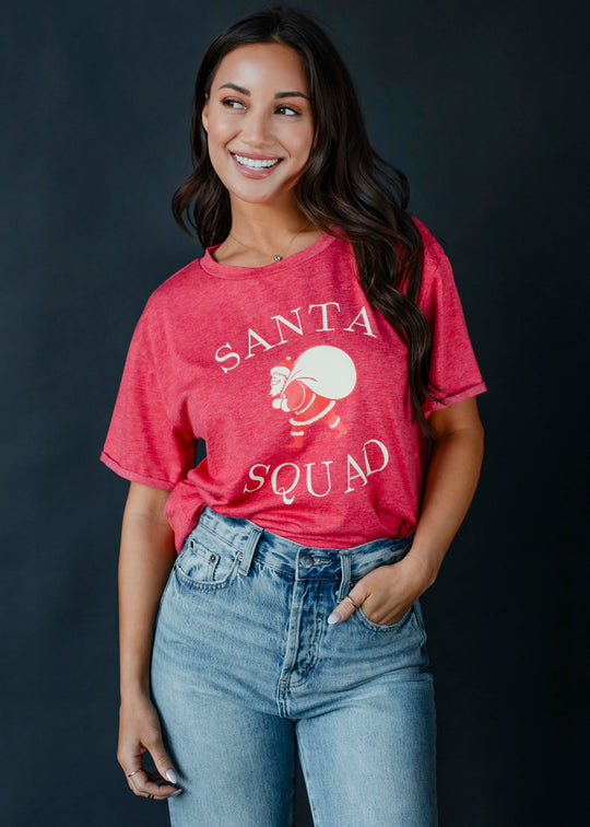 A woman with long dark hair wears the panacheaccessories Santa Squad Tee—a vintage red graphic shirt—with light blue jeans, posing against a dark backdrop and one hand in her pocket.
