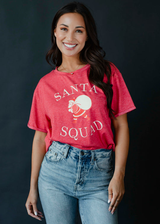 A smiling woman with long brown hair wears the panacheaccessories Santa Squad Tee, a vintage red graphic shirt, tucked into high-waisted blue jeans. She stands against a dark background, radiating festive cheer.
