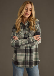A woman with long blonde hair stands against a plain background, wearing the Dakota Super Soft Button Up by panacheaccessories in green and gray plaid with front pockets, paired with light blue jeans. Her arms are crossed as she looks slightly downward.