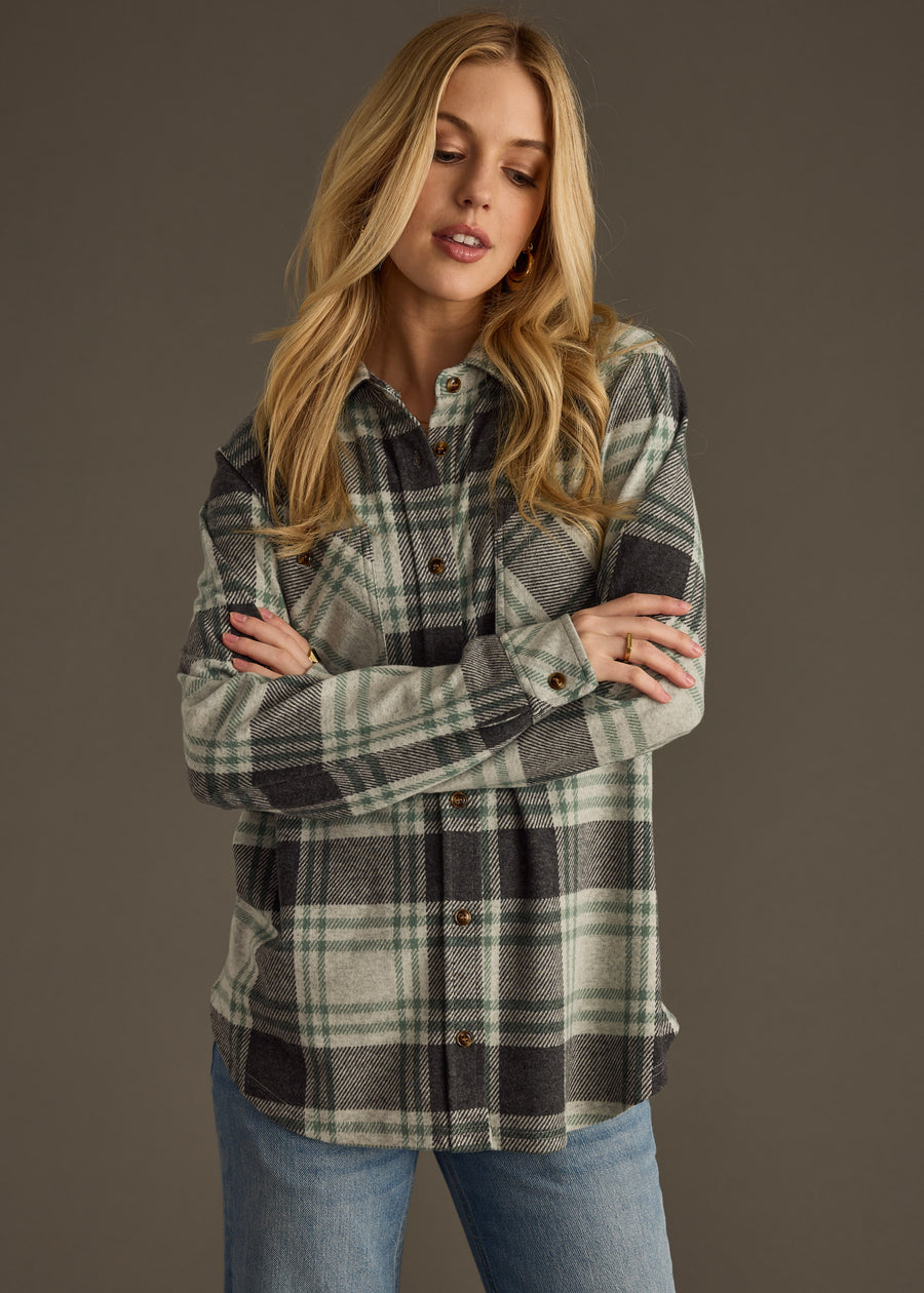 A woman with long blonde hair stands against a plain background, wearing the Dakota Super Soft Button Up by panacheaccessories in green and gray plaid with front pockets, paired with light blue jeans. Her arms are crossed as she looks slightly downward.