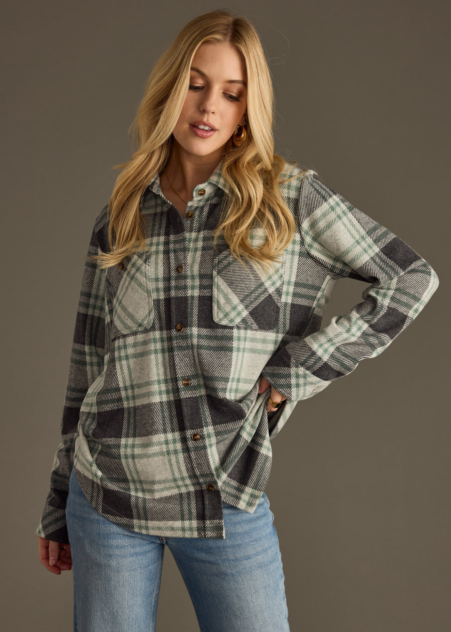 A woman with long blonde hair wears the panacheaccessories Dakota Super Soft Button Up in green and gray plaid with light blue jeans, standing against a plain gray background, one hand in her pocket and looking down.