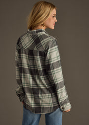 A woman with blonde hair, wearing the panacheaccessories Dakota Super Soft Button Up flannel shirt and blue jeans, stands at an angle looking over her right shoulder against a plain dark background.