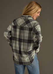 A woman with long blonde hair faces away, smiling, wearing the panacheaccessories Dakota Super Soft Button Up in green and black plaid with blue jeans against a dark background.