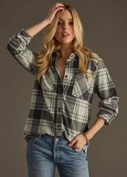 A woman with long blonde hair models the Dakota Super Soft Button Up in charcoal green plaid by panacheaccessories, paired with blue jeans, standing against a plain background with one hand on her head and one in her pocket.