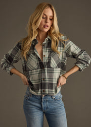 A woman with long blonde hair wears the Dakota Super Soft Button Up by panacheaccessories in green and gray plaid with light blue jeans, posing with her hands on her hips against a plain brown background.