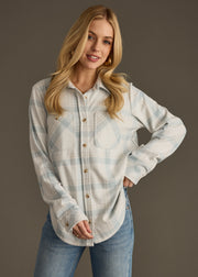 A woman with long blonde hair wears the panacheaccessories Jules Super Soft Button Up in dusty teal plaid and blue jeans, standing against a dark background, smiling with one hand on her hip.