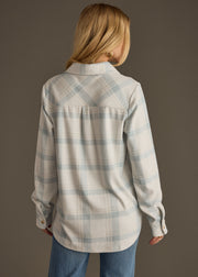 A woman with long blonde hair faces away, wearing the panacheaccessories Jules Super Soft Button Up in dusty teal plaid and blue jeans, against a plain brown background.