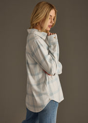 A woman with long blonde hair wears the Jules Super Soft Button Up by panacheaccessories in dusty teal plaid with blue jeans, standing sideways with crossed arms and looking slightly downward against a plain background.
