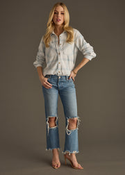 A woman with long, wavy blonde hair stands against a plain background, wearing the panacheaccessories Jules Super Soft Button Up in dusty teal plaid, ripped blue jeans with knee holes, and nude heels.