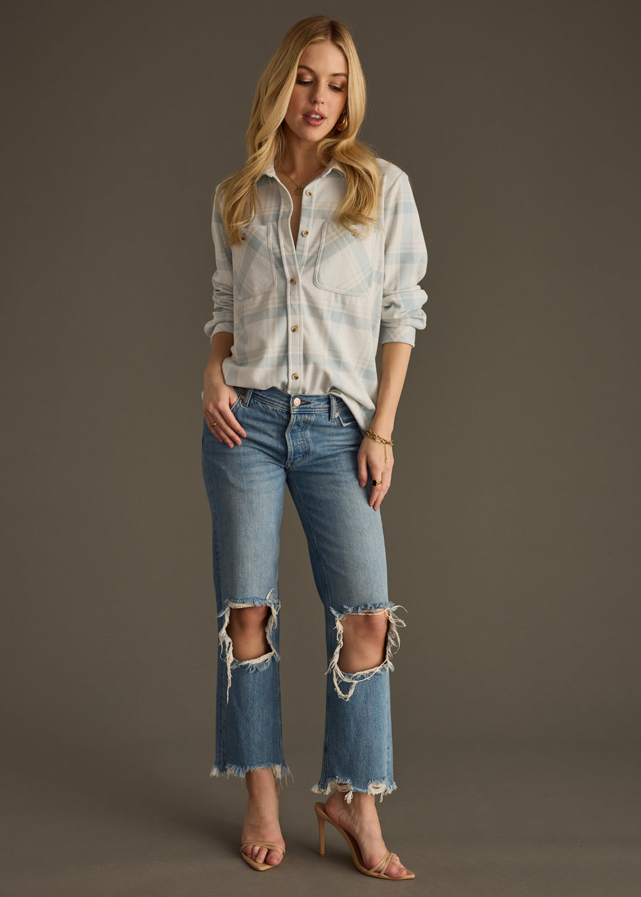 A woman with long blonde hair poses against a plain background wearing panacheaccessories’ Jules Super Soft Button Up in dusty teal plaid, paired with ripped blue jeans and nude high-heeled sandals.
