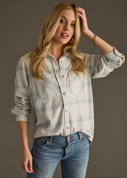 A woman with long blonde hair wears the Jules Super Soft Button Up by panacheaccessories in dusty teal plaid and blue jeans, standing against a plain background with one hand on her head and the other at her side.