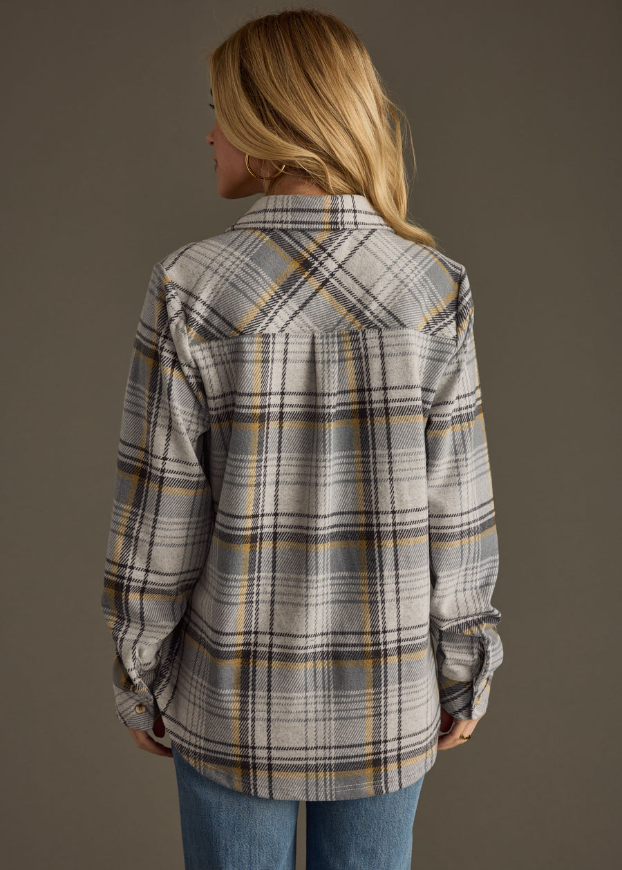 A woman with blonde hair, seen from behind, wears the Ozzy Super Soft Button Up by panacheaccessories in gray, white, and yellow plaid with blue jeans against a plain background.