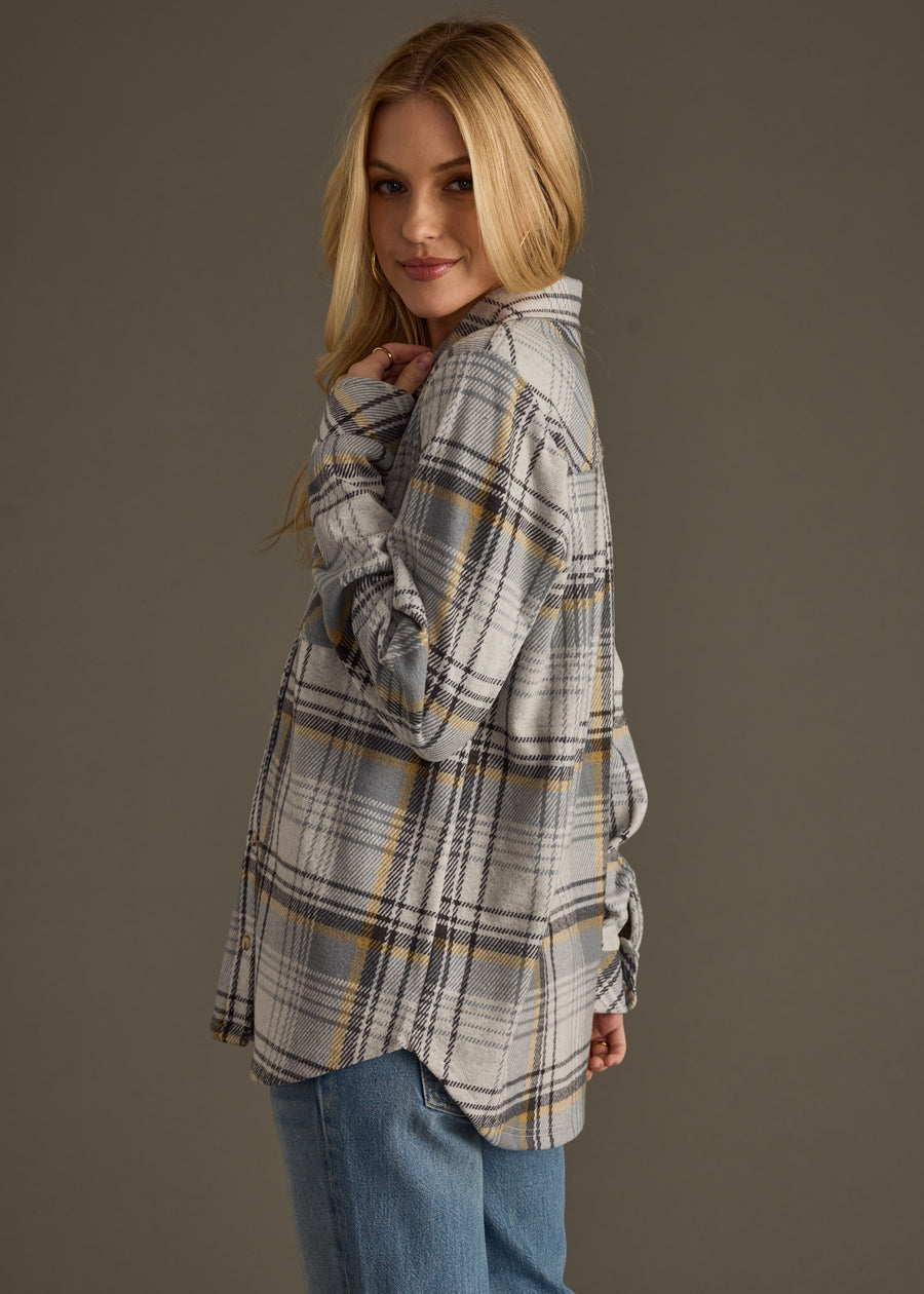 A woman with long blonde hair wears the panacheaccessories Ozzy Super Soft Button Up in grey plaid with blue jeans, standing sideways and smiling over her shoulder against a plain background.