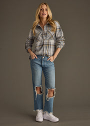 A woman with long blonde hair and a relaxed expression stands against a plain background, wearing the panacheaccessories Ozzy Super Soft Button Up in gray plaid, ripped blue jeans, and white sneakers with her hands in her pockets.
