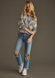 A woman with long blonde hair stands against a plain background wearing the Ozzy Super Soft Button Up by panacheaccessories, paired with ripped blue jeans and white sneakers. She looks down, holding one wrist, showcasing the cozy gray plaid shirt.