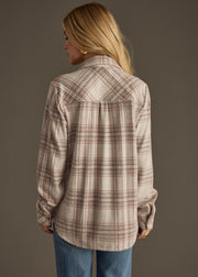 A person with long blonde hair, wearing the panacheaccessories Lia Super Soft Button Up in beige and pink plaid and blue jeans, stands facing away from the camera against a plain brown background.