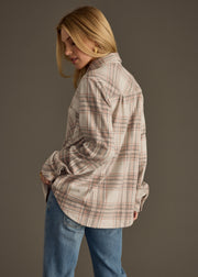 A woman with blonde hair wears the Lia Super Soft Button Up by panacheaccessories—a beige and pink plaid shirt—paired with blue jeans. She stands in front of a plain background, turned slightly to the side and looking down.