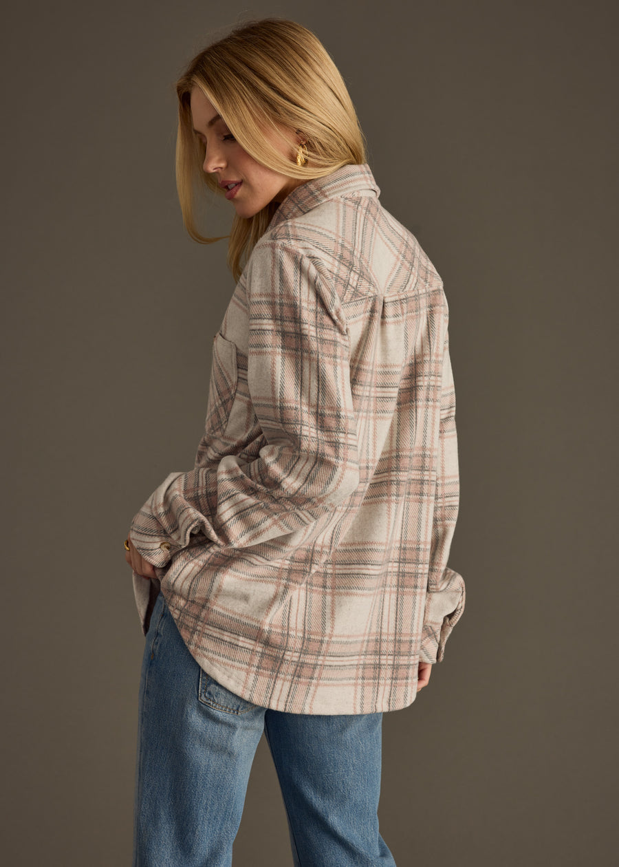 A woman with blonde hair wears the Lia Super Soft Button Up by panacheaccessories—a beige and pink plaid shirt—paired with blue jeans. She stands in front of a plain background, turned slightly to the side and looking down.