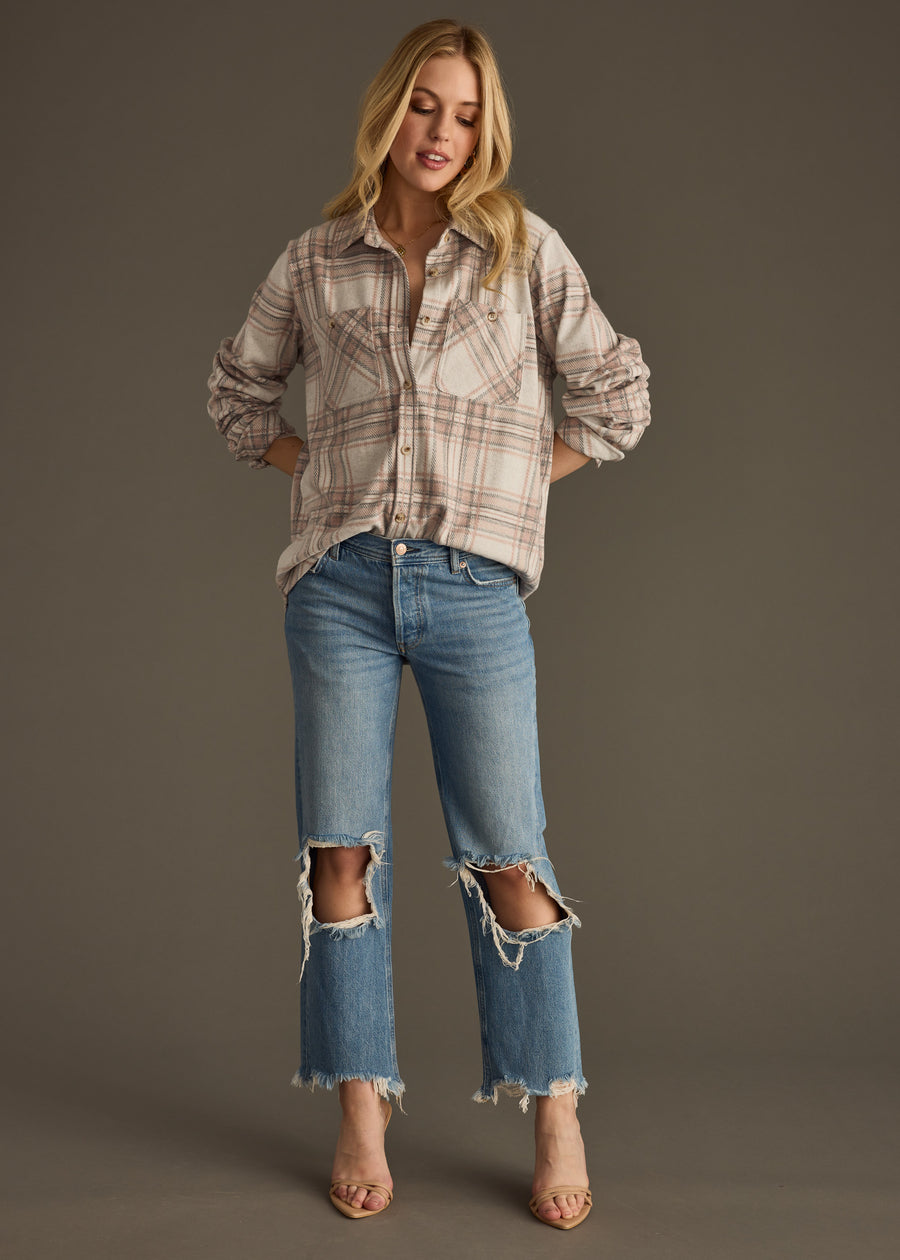 A woman with long blonde hair stands against a plain background wearing panacheaccessories’ Lia Super Soft Button Up and ripped blue jeans with frayed hems. She has open-toe heels, hands in her pockets, and a relaxed expression.