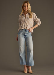 A woman poses confidently with one hand in her pocket, wearing the panacheaccessories Hilarie Super Soft Button Up in beige plaid, paired with light blue high-waisted jeans and nude heels against a plain background.