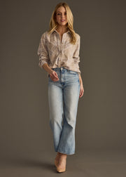 A woman with long blonde hair stands against a plain background wearing the Hilarie Super Soft Button Up by panacheaccessories, light blue high-waisted jeans, and beige heels. She gazes past the camera with a relaxed expression.