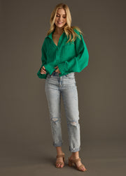 A woman smiles against a plain background, wearing the Wild Heart Button Up - Green from panacheaccessories, styled with light-wash ripped jeans and tan heeled sandals.