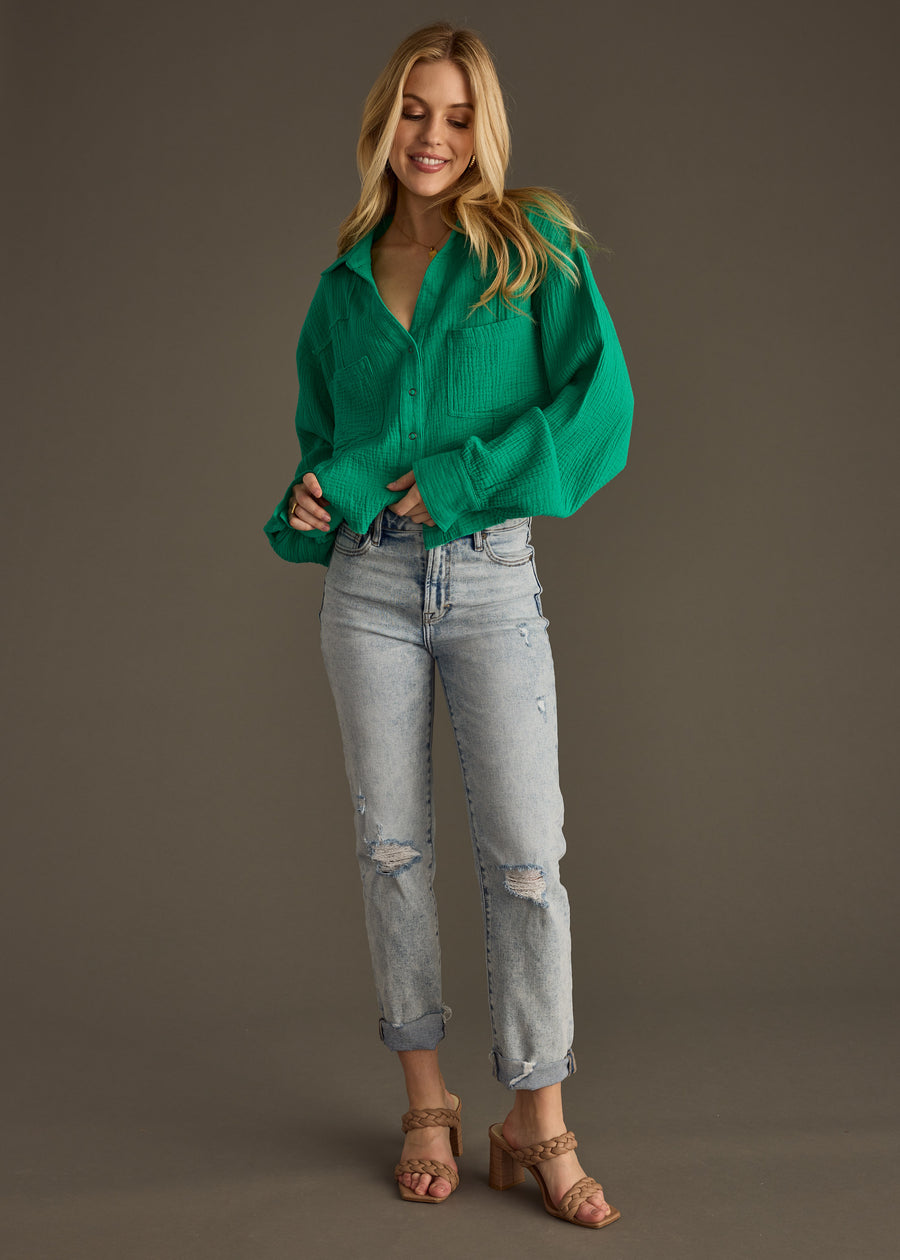 A woman smiles against a plain background, wearing the Wild Heart Button Up - Green from panacheaccessories, styled with light-wash ripped jeans and tan heeled sandals.