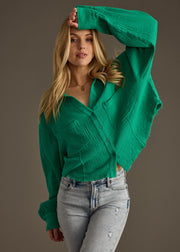 A woman with long blonde hair poses with one arm raised, wearing the panacheaccessories Wild Heart Button Up in green and light-wash ripped jeans against a plain gray background.